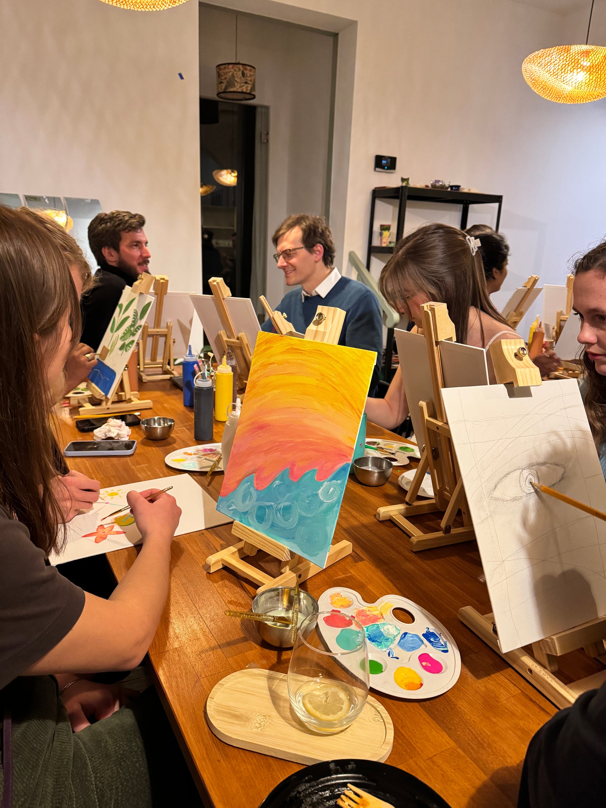 A group of people seated around a table at an art and craft session, with easels and paint trays in front of them.