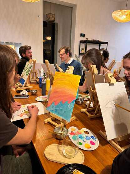 A group of people seated around a table at an art and craft session, with easels and paint trays in front of them.