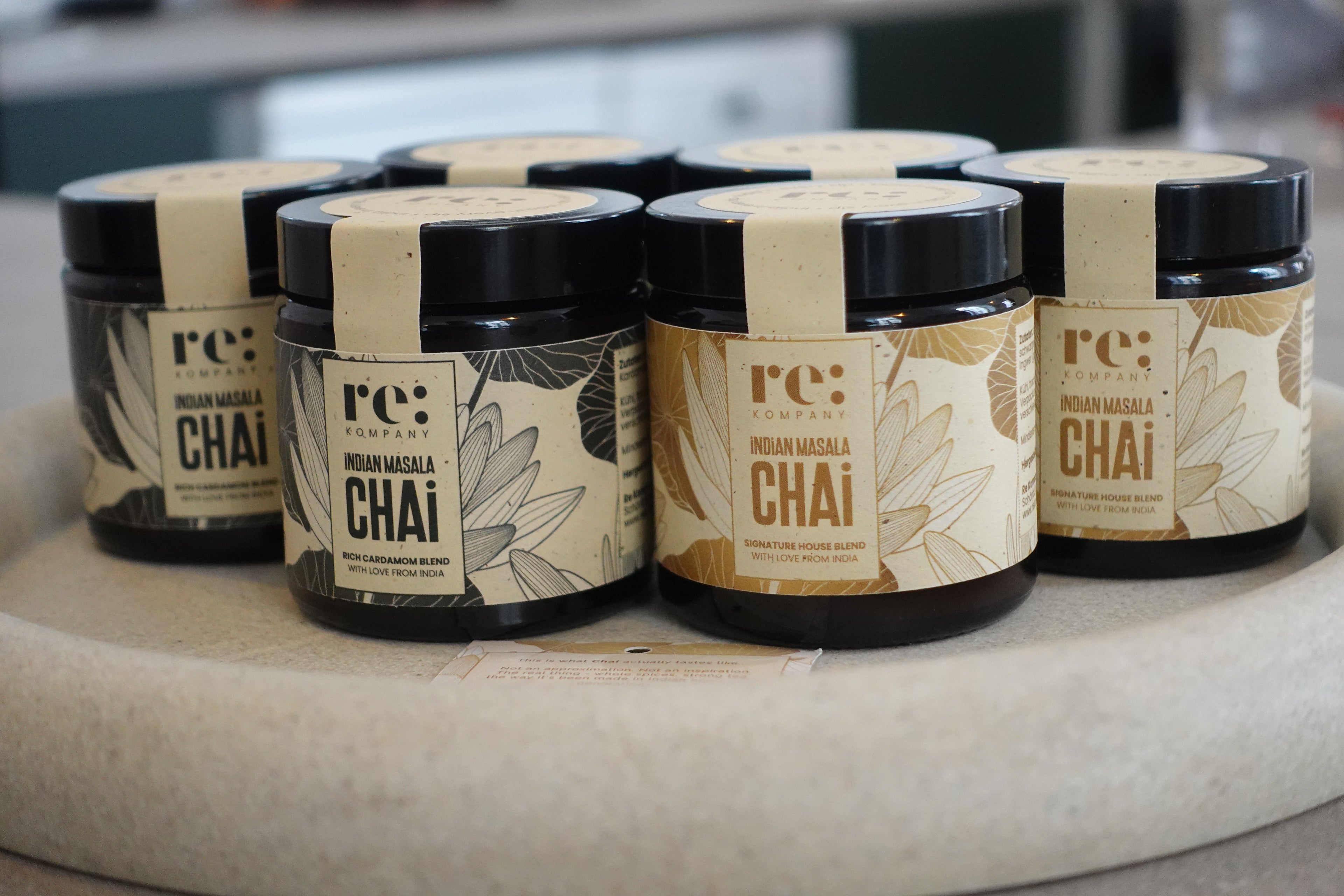 Six glass jars of rekompany's Indian Masala Chai arranged on a round stone tray. The jars have black lids and labels with beige and brown botanical designs, with one jar showing a dark chocolate-colored chai powder inside.