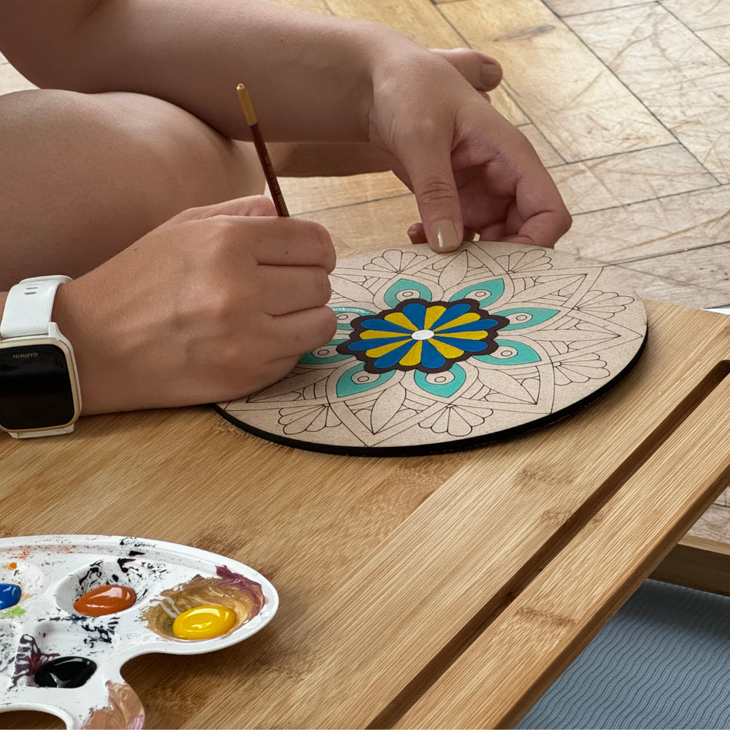 Person painting a mandala on a wooden board with a paintbrush and palette on a wooden table.