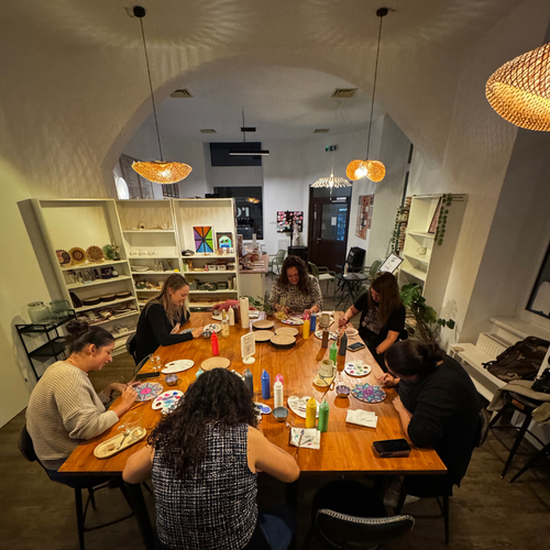 Group of people sitting around a wooden table, painting Mandalas in a modern cafe with shelves and decor in the background.