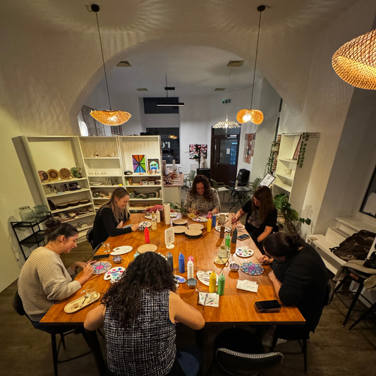 Group of people sitting around a wooden table, painting Mandalas in a modern cafe with shelves and decor in the background.