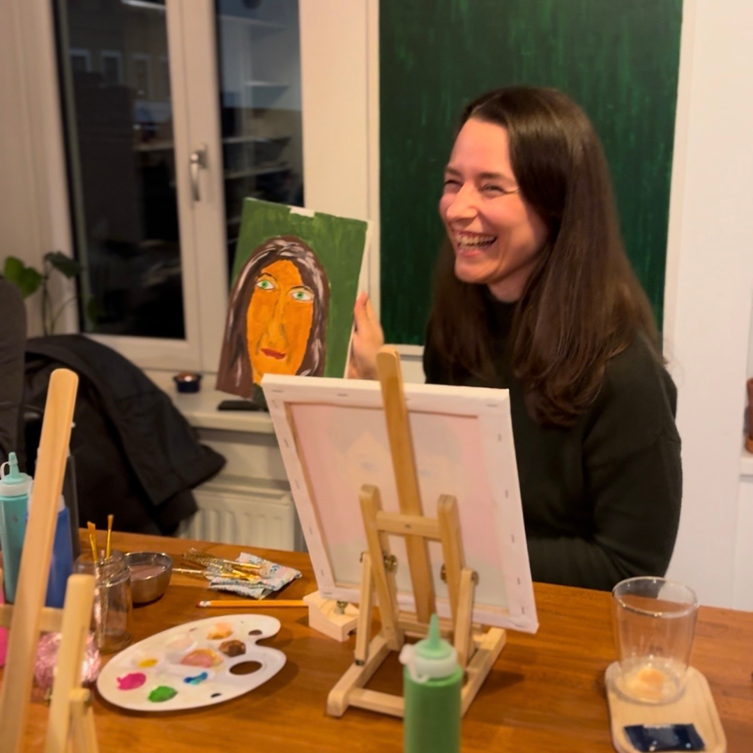 Woman sitting at a table with art supplies, including a painting and easel, in an indoor setting.